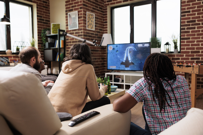 Multiethnic group of friends sitting on sofa at home while playing space videogame on gaming console.